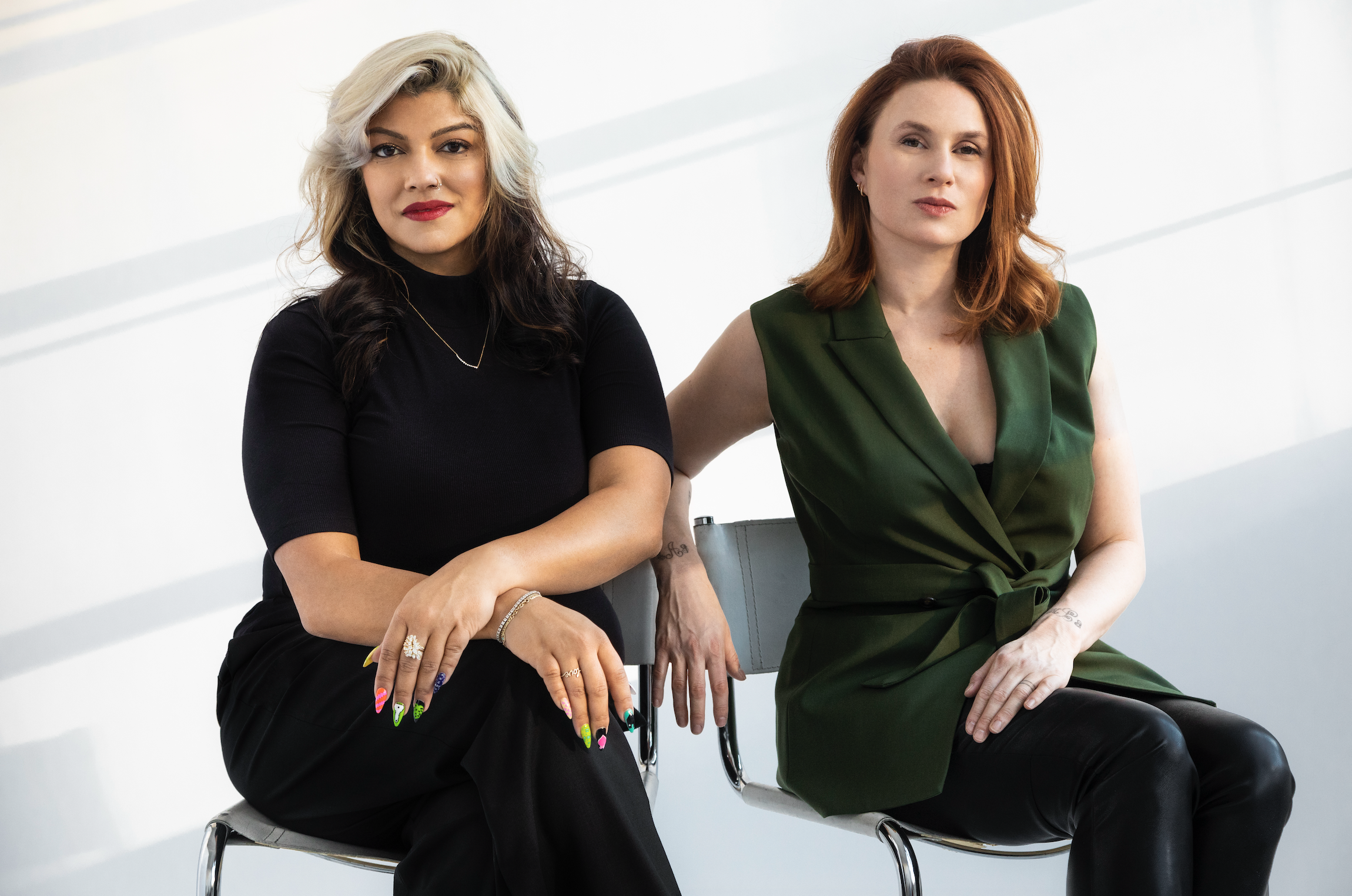 Portrait of two women sitting on metal chairs against a neutral white background.